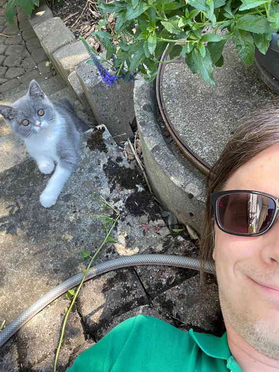 On the left side we see Spacy the kitten from my parents and on the right side half of my face is visible (smiling and wearing sunglasses and a green polo shirt). I lay outside on the ground. We both look into the camera.