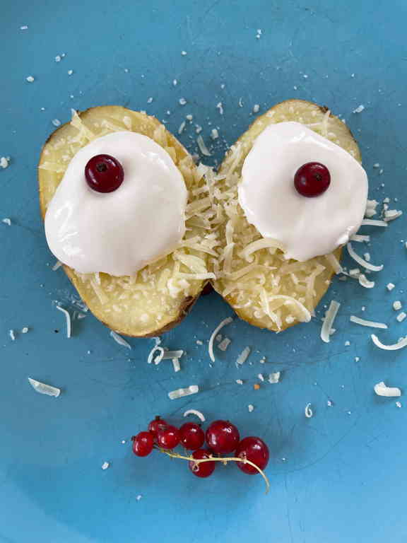 A close crop of a face made from two potato halves with sour cream on top for eyes and a single currant for the pupils and multiple currants for the mouth. The face has a slight panic look to it (from the position of the pupils).