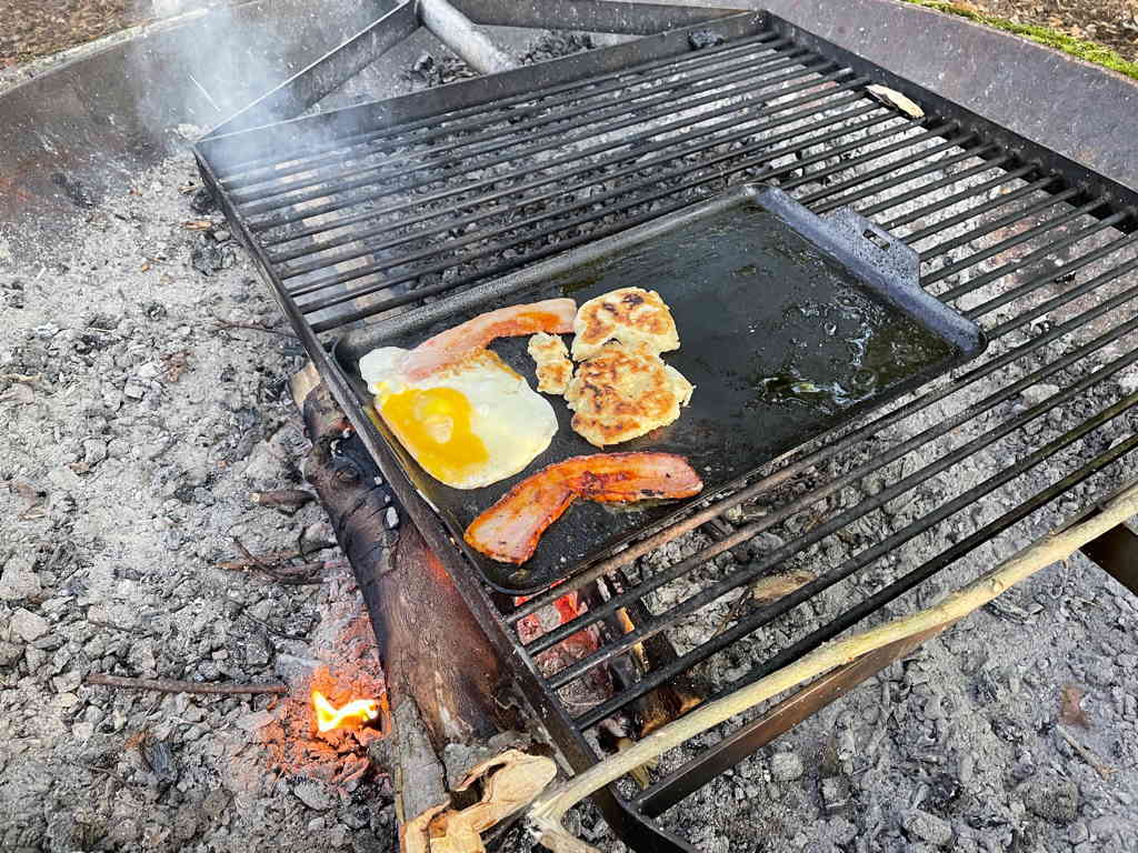 Da close shot of a fire with a grill on it. An egg and some bacon are freeing in a pan on the grill.