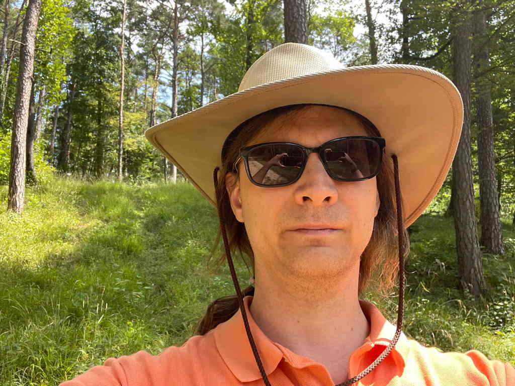Zoomed-in selfie of me in the forest. I wear a “cowboy” inspired hat and sunglasses. At the bottom part of my orange polo shirt is visible. In the background you see the sun flooded forest. It is a light forest with a lot of grass at the bottom.