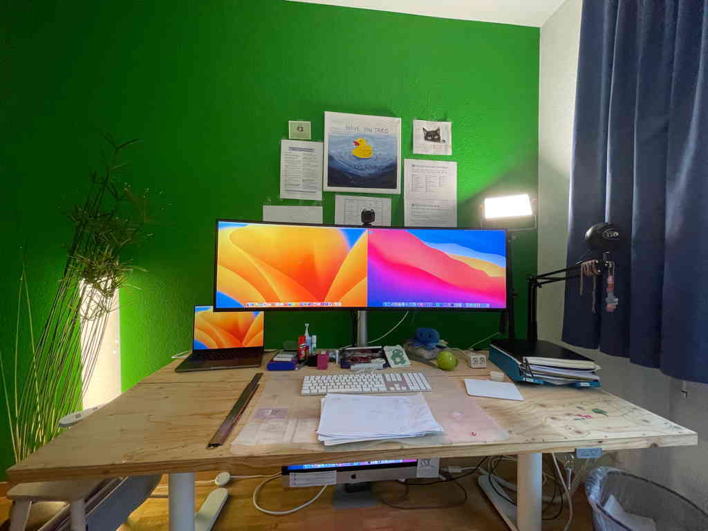 In the center the new computer screen is visible, showing the desktops of two Macs. On the left the new Laptop for work and right my old iMac. The desk is freshly cleaned. In the background is a green wall. And a plant on the left side of the desk.
