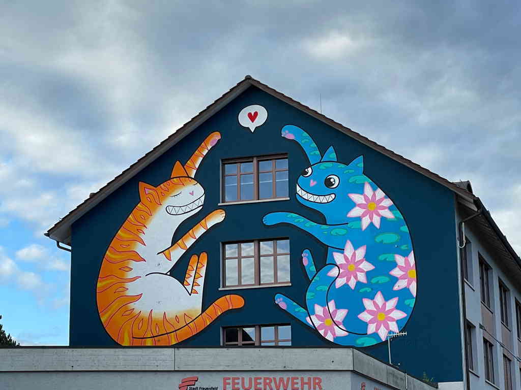 Close-up of a house side with two cats drawn on it. The left at is orange the right cat is blue with decorative flowers and a big grin.