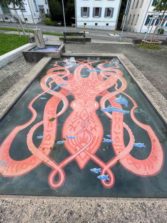 A zoomed in picture of a fountain. Inside the fountain there is a picture of a squid filling the whole floor of the fountain. Some blue fish are drawn on top of it as well.
