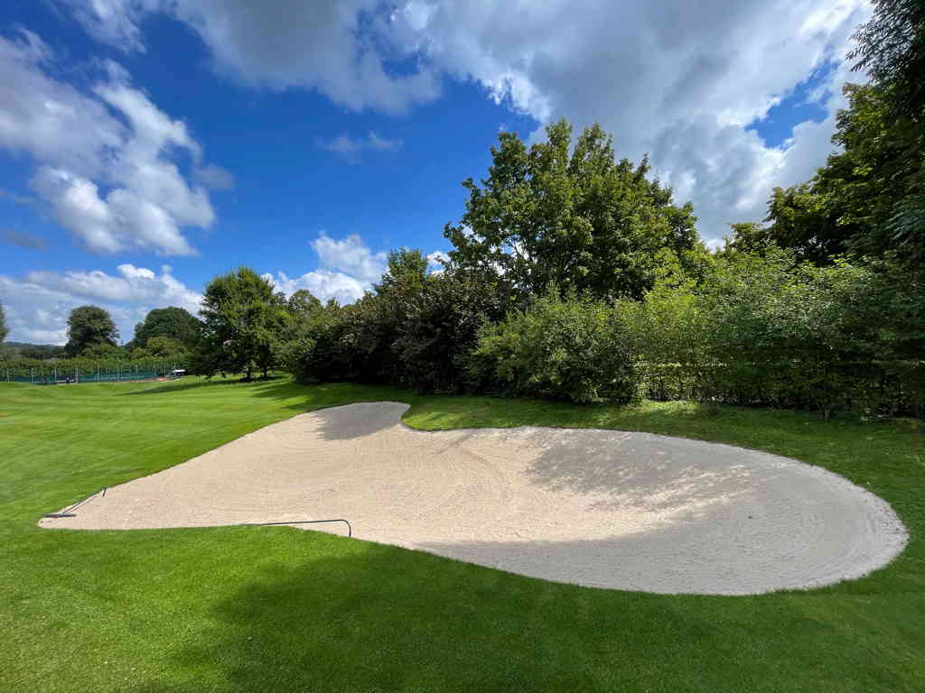 A wide shot of a golf bunker in the shape of a heart. On the right side (at the top of the heart) are some trees creating shadows. And the sky is filled with big clouds, but some blue is still visible.