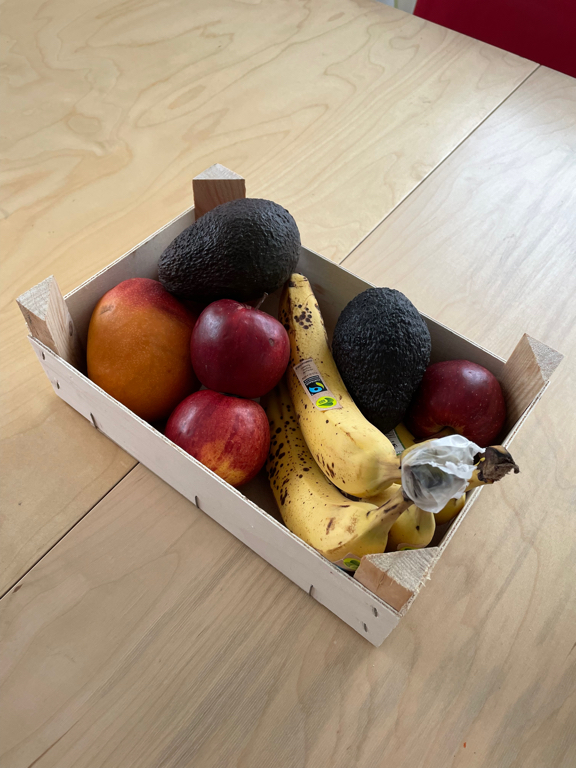 A wooden crate with fruits. It has bananas, avocados, apples and a mango in it. 