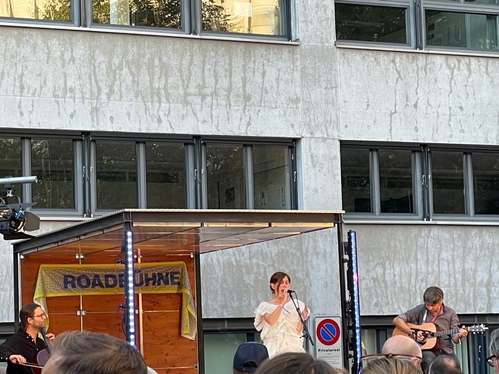 In the center the singer is singing. She wears a white dress. On the left side is a cello player and on the right side a guitar player. The stage has a roof and two light. In the background is grey office building.