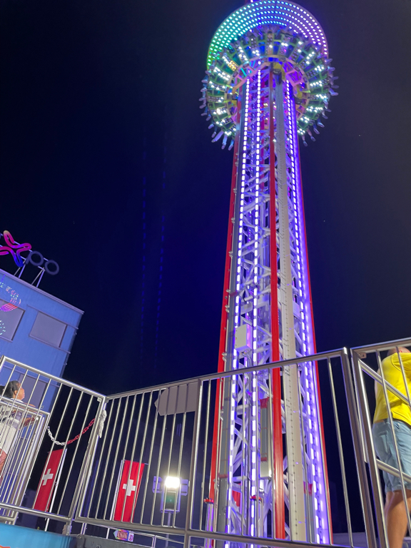 In the center of the image is one of the attraction at the Badenfahr fair. A freefall tower. It is illuminated with colorful lights. The picture is taken from the base of the tower. The ride is just falling down with a group of revelers.