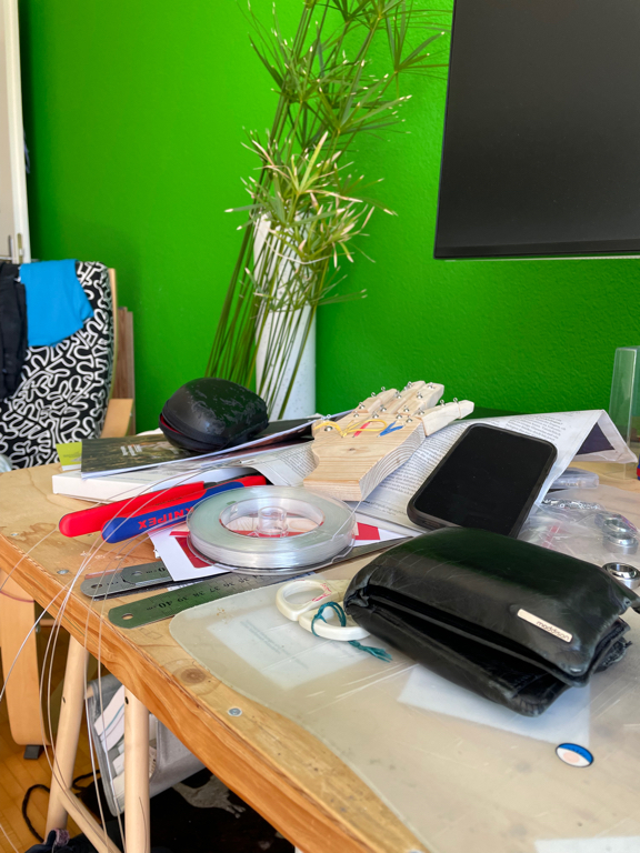A zoomed-in picture of the left side of my desk with a build up of different items: a folded open newspaper I should read, the cover of my sunglasses, my work phone. A wrench and parts of my current wood working project. In the foreground is also my wallet. 