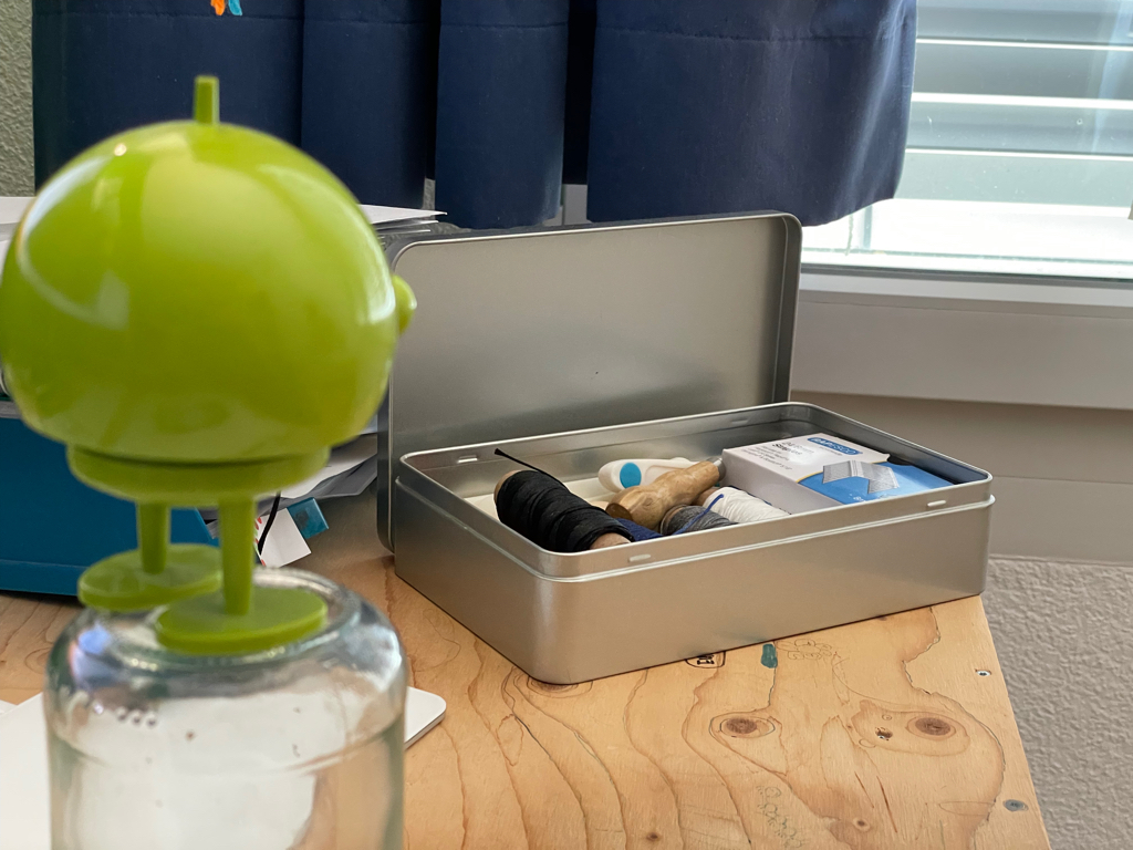 A closeup of a green Hoptimist facing away from the camera looking to the right. On the right side in the background is a big tin box filled with bookbinders yarn and implements. 