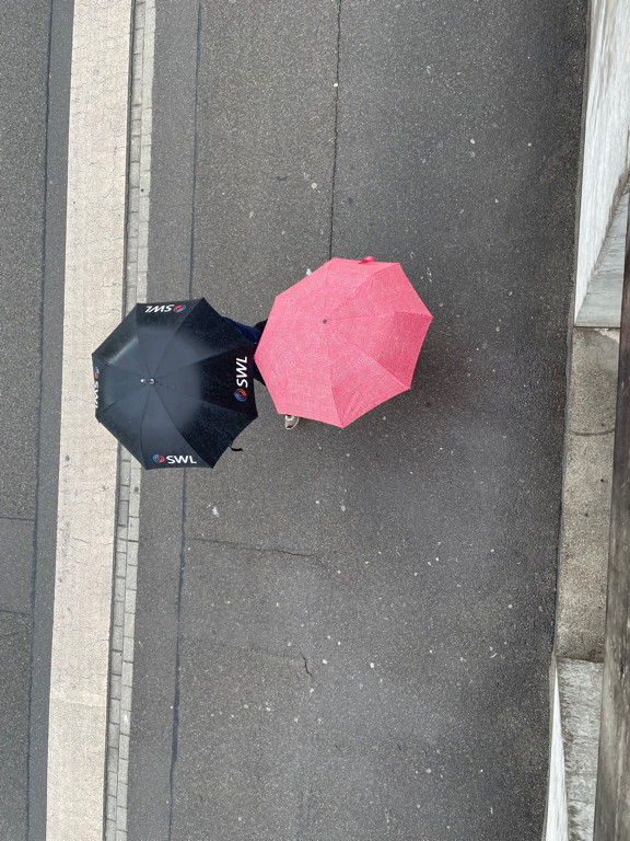 Photo of two humans beneath umbrellas. The photo was taken from a high vantage point downward. The subjects stand on concrete. One Umbrella is black, the other is pink.