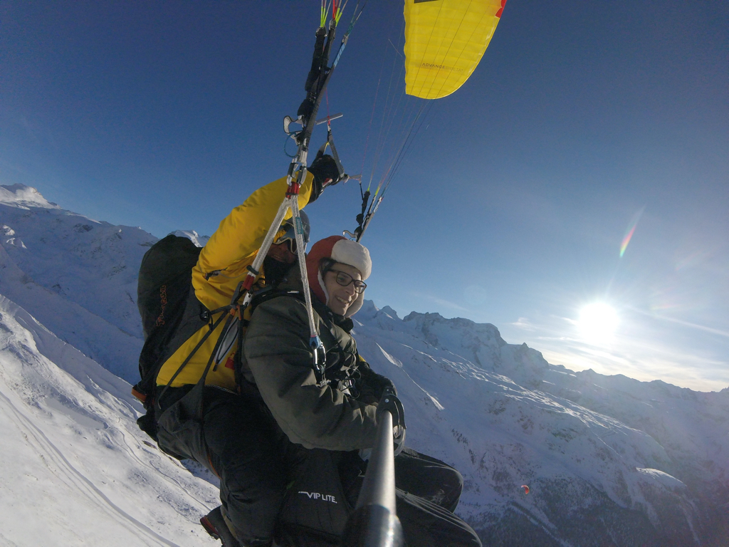 Photo of myself and the paragliding pilot shortly after takeoff. On the right the sun is thing brightly. It is winter time we are both wearing multiple layers of clothes against the cold. The background is filled with the mountains full of snow.