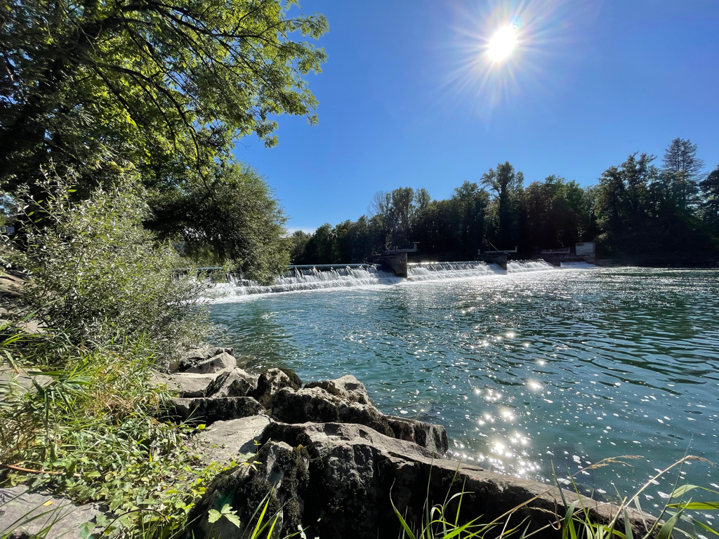 A wide shot of a weir in a river. The weir is only around 2m high. On the left side and part of the bottom the shore is visible and in the top right corner there is the bright sun shining. With some forest below in the background. The water is reflecting the bright sun and the sky is blue.