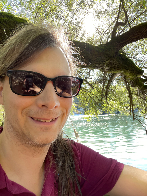 A selfie under a tree with a river in the background. I’m wearing my sunglasses. And a violet polo shirt.