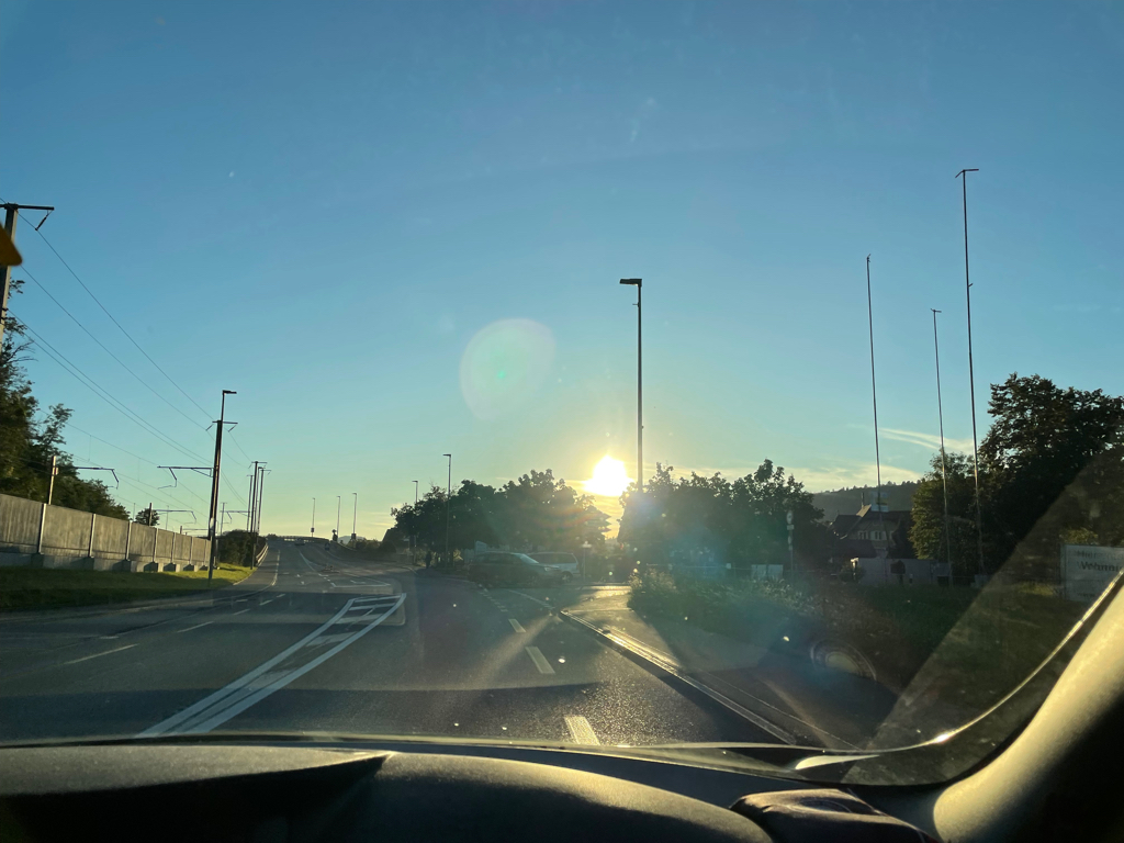 A image of the sunset photographed form the car. You see the sun setting behind some trees. And part of the dashboard ob the car at the bottom border of the image.