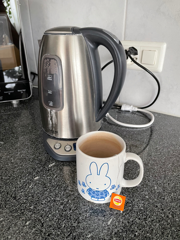 The image shows a close-up of our water cooker with a mug in front of it. The mug contains a tea-bag (you only see the string of it and its tag). The mug has the image of Nijntje (Miffy) on it.
