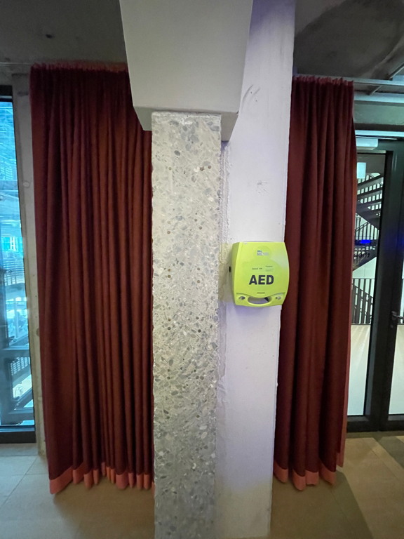 A photo of a concrete pillar with two dark red curtains next to it. A bright green  AED device hangs in the middle and to the right of the pillar. The concrete has a cut and polished look and you see the exposed aggregate in it.