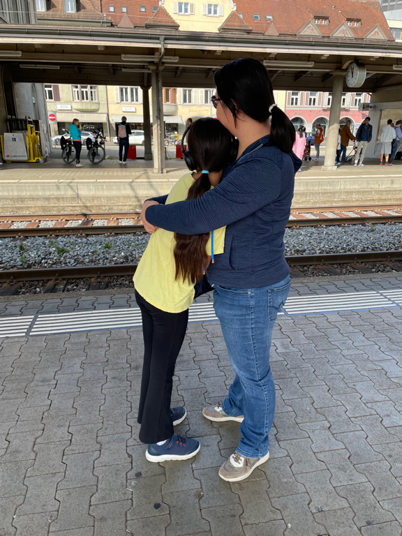 An image of my wife and daughter embracing each other. Both look away from the camera. They stand at a railwai station and in the background other travelers are visible. My daughter wears a yellow shirt and black pants. My wife a blue jacket and jeans. 