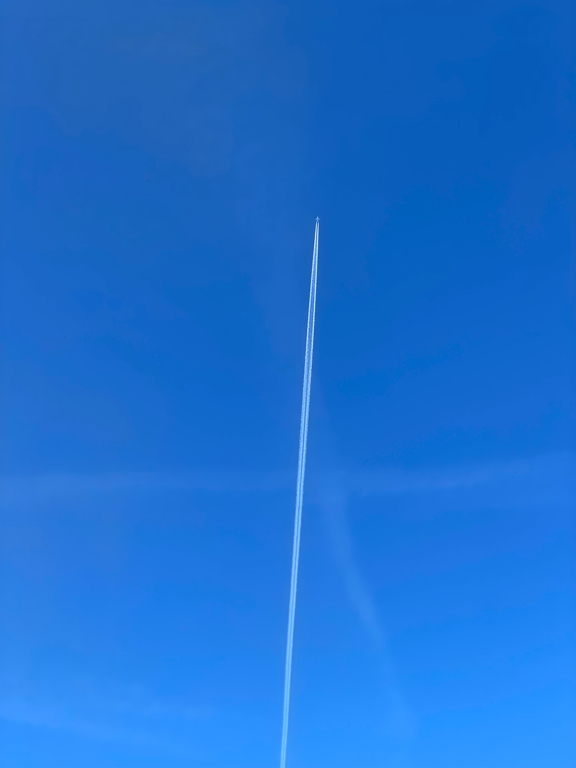 A wide shot of the blue sky with a single contrail in the middle, and the airplane at the end of it. The contrail goes from the bottom edge to around two thirds of the image height. There are some older and fainter contrails in the background as well, making a faint cross.
