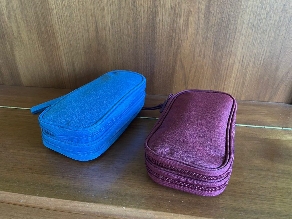 Close-up picture of two fabric cases for cables and power bricks. The one on the left is blue the other one is burgundy red. Photographed on a wooden background.
