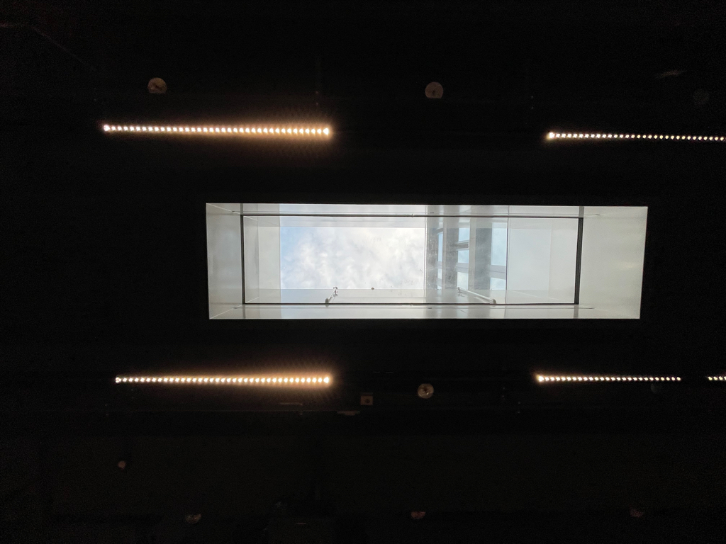 A picture of the ceiling with a skylight in the middle. Everything except for the skylight is in the shadows. The skylight is surrounded by four lights. 