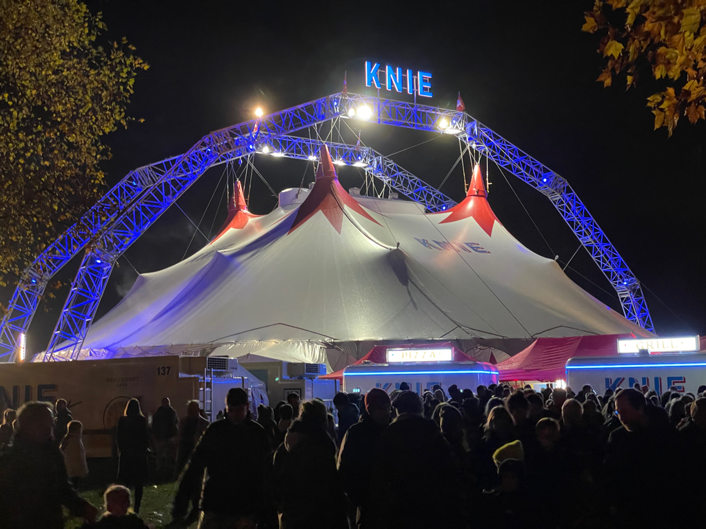 Picture of the circus tent from the outside. It is night time and the tent is illuminated. The truss holding up the ten fabric is blue and has an illuminated logo reading “Knie” on it. In the foreground a crowd of people are visible.