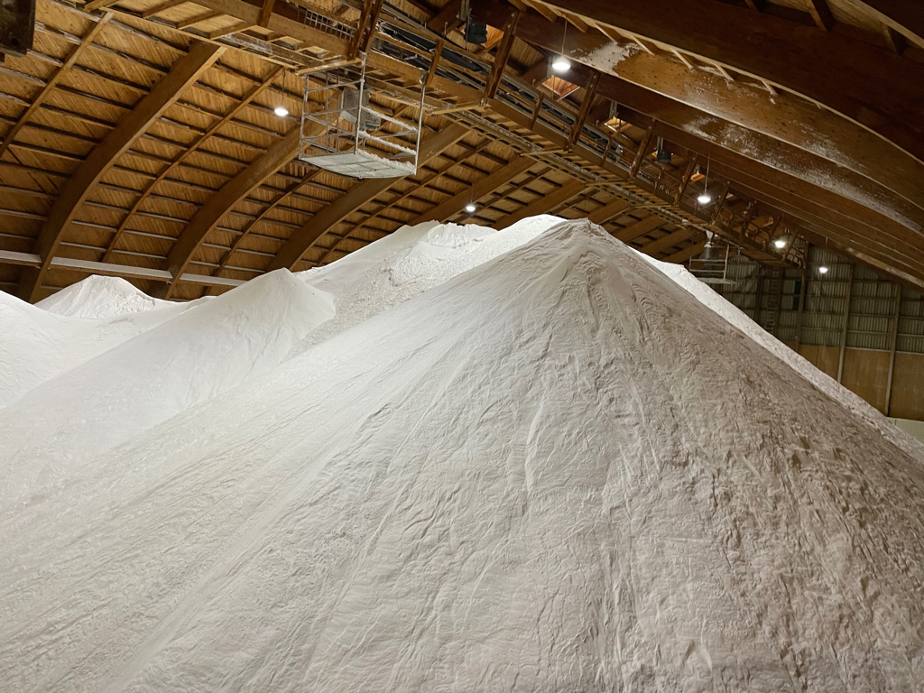 A closeup of a salt mountain inside a wood shed. The mountain has multiple tops and you see mostly salt. At the top in the middle is conveyor belt to add more salt.