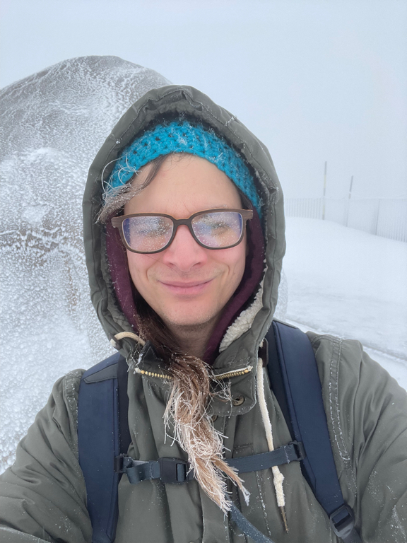 A selfie of myself on top the mountain in the fog and snow. The wind is driving the snow into my face and I have my eyes half closed behind my glasses.