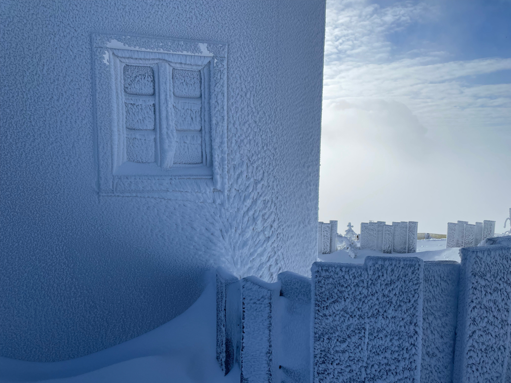The photo shows the corner of a house covered in hoarefrost. The frost forms a pattern looking like a tree growing up the side of the house. In the freground is a wall covering part of the house wall. And on the right side you see the clouds rising up from the valley again.