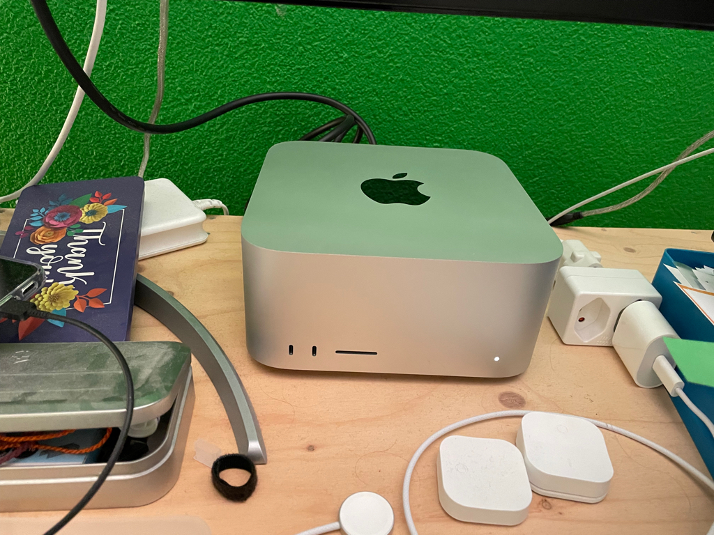 A close-up photo of a Mac Studio on my desk. Around it there are cables and office stuff. The Mac stands in front of a green wall.
