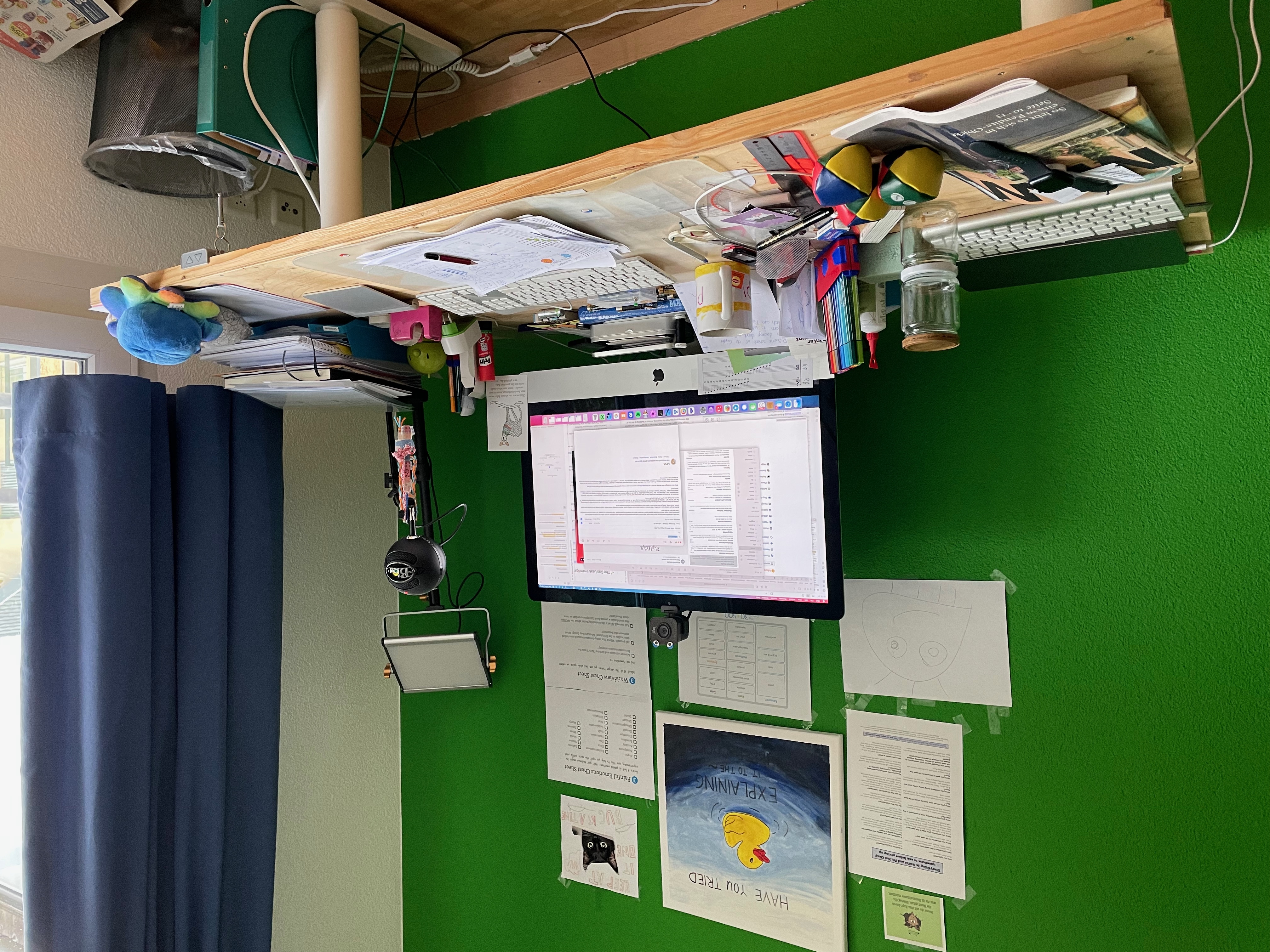Unscensored image of my desk. It is filled with stuff. There are a MacBook pro and an iMac on it, my jougling balls my stack of papers and drawing utensils. A collection of old batteries.