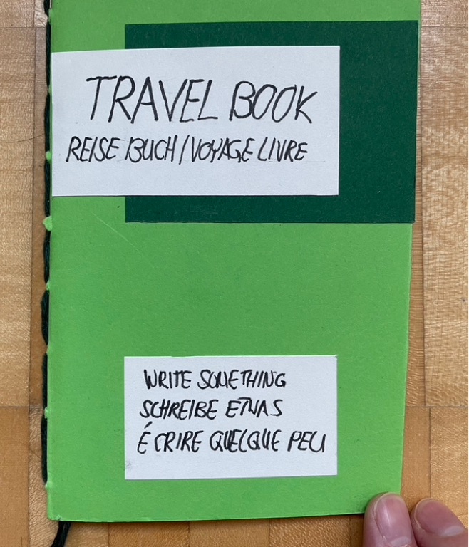 Text in three languages: Travel Book and the same title in German and French, as well as the prompt “Write Something” also in these three languages. The cover is green and the text is on a white background which was glued on top of the cover.