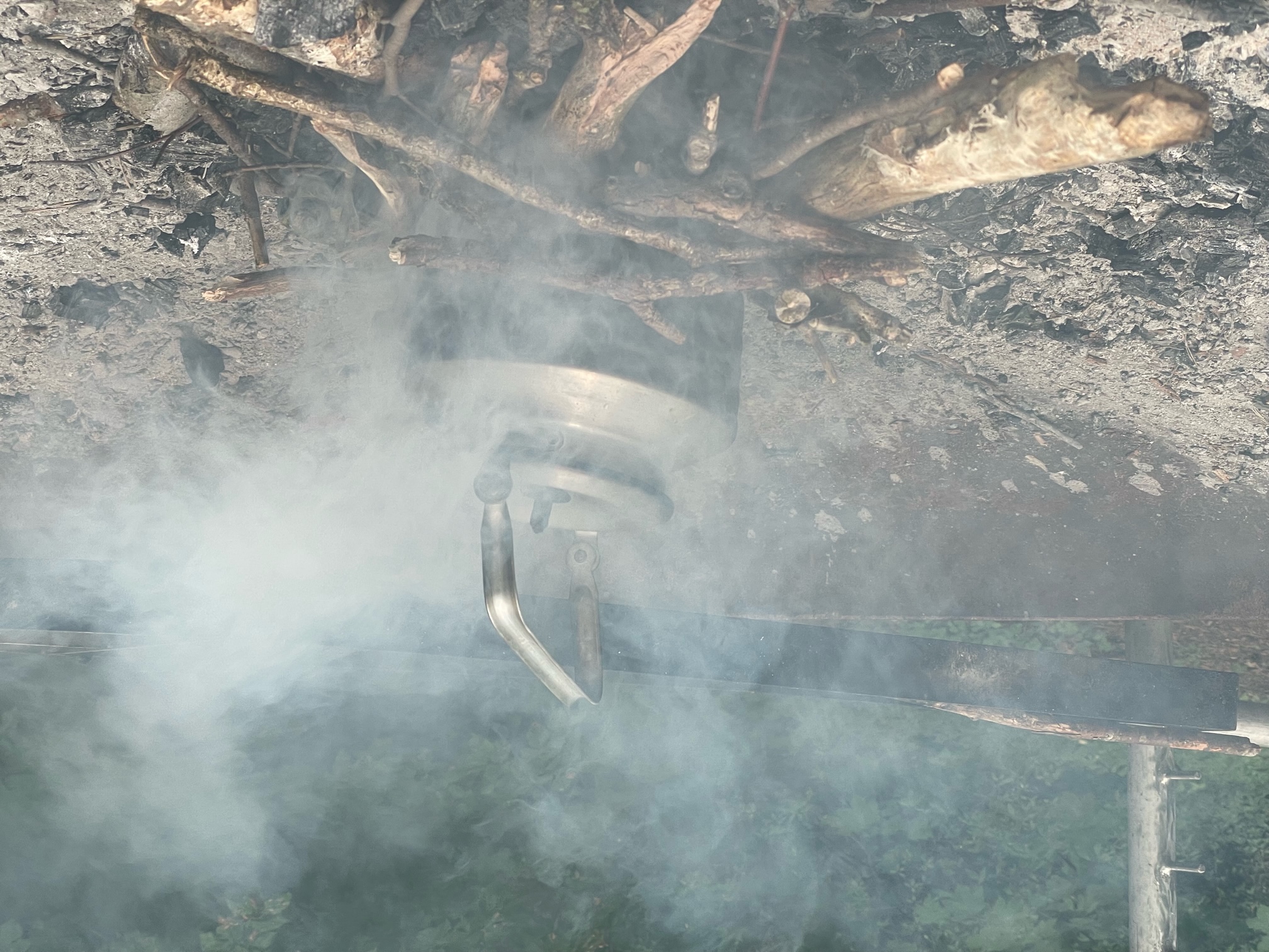 A fire outside without a visible flame and a lot of smoke. A tea kettle is on the top.