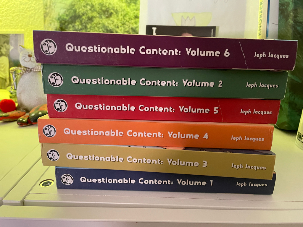 A close-up of a stack of books lying on the side. The spine is facing the camera. It is book one to six of the webcomic questionable content.