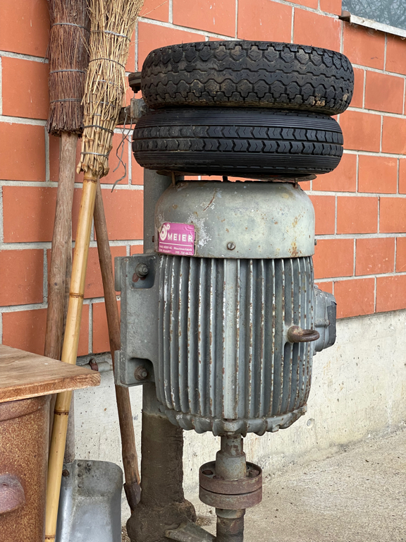 A electrical motor installed vertically, with the axis pointing down. On top there are two tires to protect the motor from being bumped into. It is a big (at least 50cm long) greenish industrial looking motor.