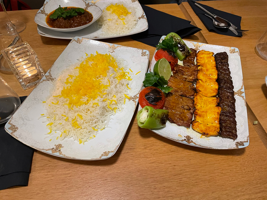 Two plates with food on a table. The left plate is filled with rice (with some yellow stuff on top) and the right plate contains (from left to right): A row of vegetables a row of Lamb fillet, a row of chicken filet and in the last row we have Kabab koobideh.
