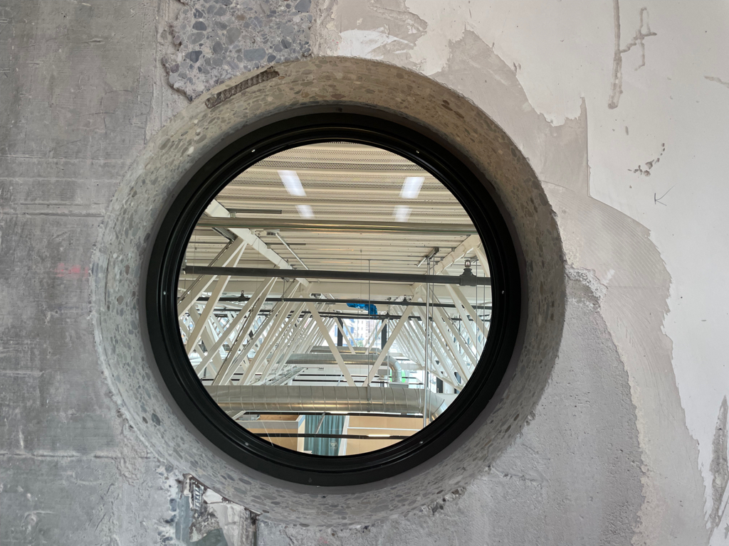 A round window in a concrete wall. The window has a black border. Through the window, you see the metal trusses of the roof of the next room. 