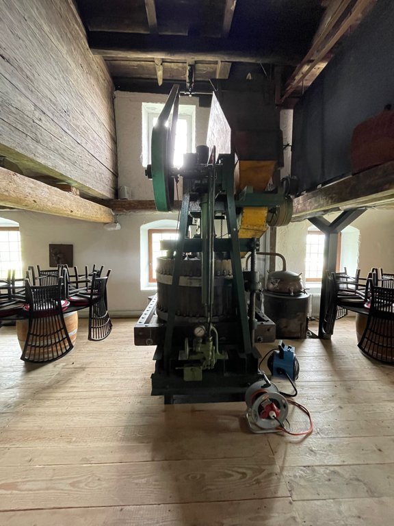 An old apple juice press. In a semi-dark room. Behind the machine are some windows casting the machine in harsh light. 
