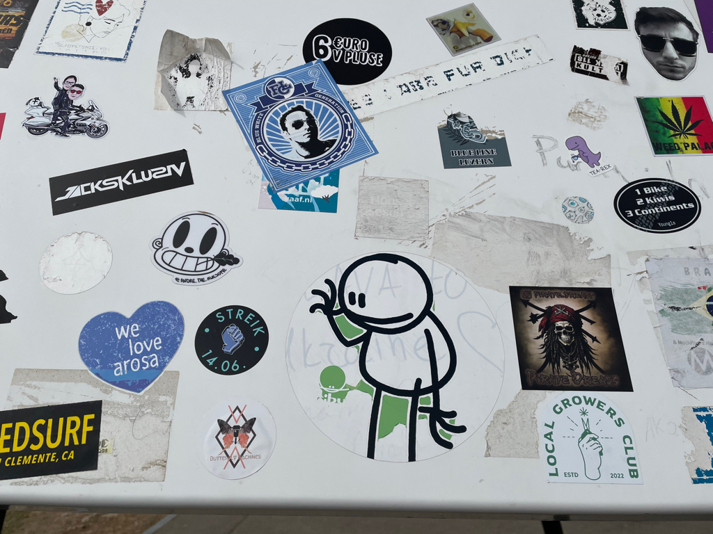 A close-up of a wall of different stickers. The main focus of the image is a sticker of a waving stick figure. 