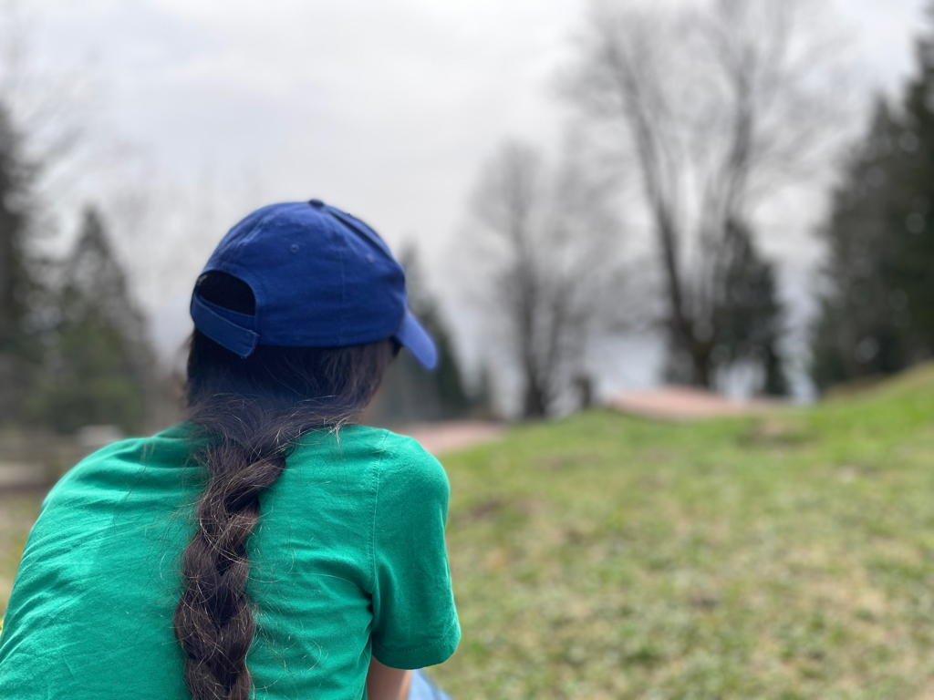 A girl photographed from behind on the left side of the image. She is wearing a green T-Shirt and a blue cap. The hair is braided. The background is blurred and showed trees and grass. 