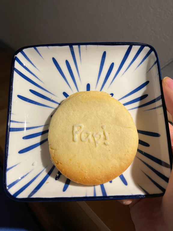 A round cookie on a plate. The cookie is decorated with white sugar frosting spelling the word “Papi” (dad in German). The cookie is on a white board with blue stripes in a radial pattern toward the centre - where the cookie lies. The pate is hold by a hand on the right side part of the hand holding it is just visible.
