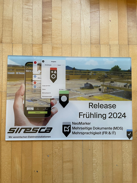 A poster printer behind a sheet of acryl glass. On the left side is a visualisation showing a hand holding a phone with our app running – showing the primary new feature of the app. Markers which can indicate information on a plan with different properties. The left side has the title “Release Frühling 2024” and below is a list reading: NeoMarker, Mehrseitige Dokumente (MDS) and Mehrsprachigkeit (FR & IT). At the bottom left side is the company logo and our claim in German, reading: Siresca / Wir vereinfachen Elektroinstallationen (We simplify electric installations).