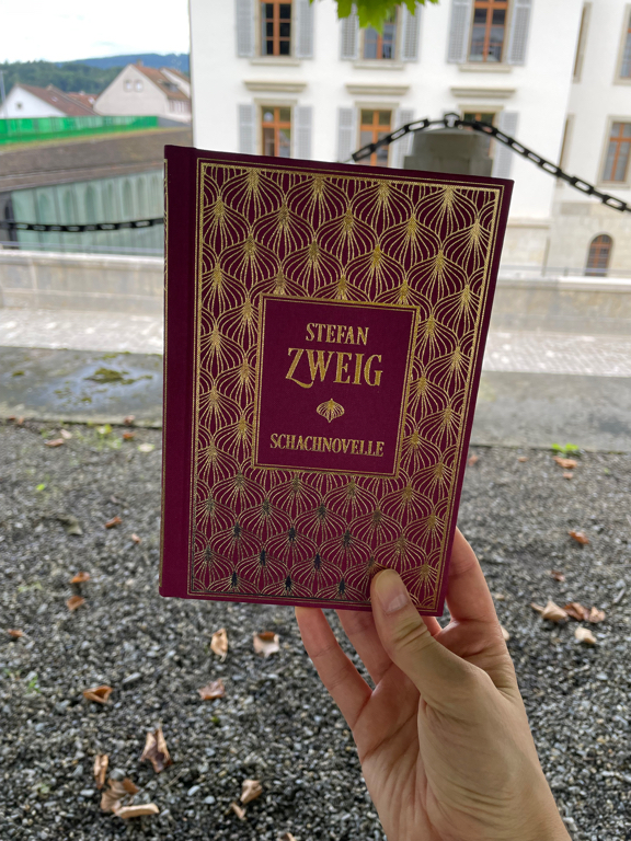 A hardcover book hold on the bottom right corner. The text reads “Stefan Zweig” and below it on the se ond line “Schachnovelle”. The cover has golden foil decoration on a dark violet background.