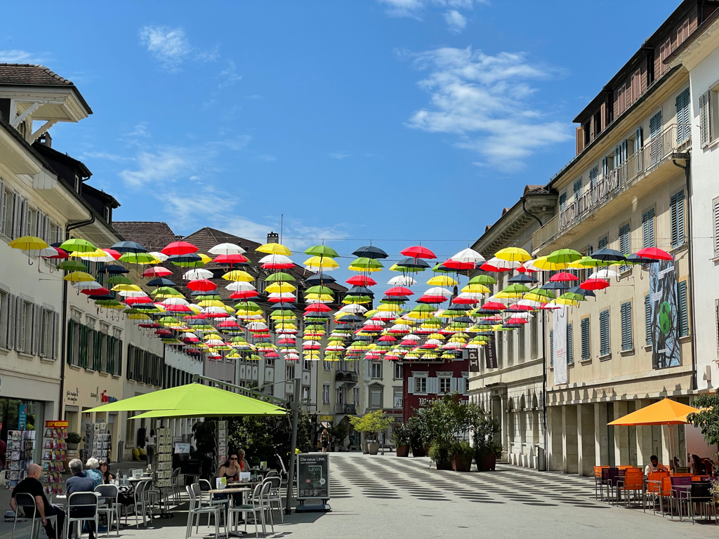 A wide citystreet withstrings of umbrellas above it. The umbrellas are in multiple colors (red, yellow, and green). Below are stores and coffee shops.