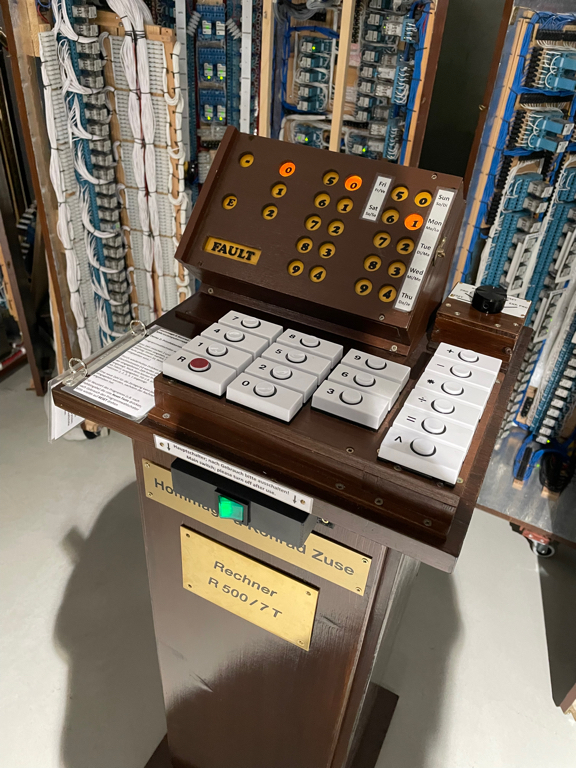 A close-up of the control panel of a relay computer. The panel is on a pedestal and the top part shows the output of the computer via multiple lamps with numbers on it. The bottom part contains multiple buttons for the input (also with numbers on it). On the right side there is also a rotary switch to choose the program to execute. In the front below the input buttons is the power button for the computer. Followed by two plaques reading Hommage Konrad Zuse (homage for Konrad Zuse) the text is partially hidden by the input panel on top. The second plaque reads Rechner (newline) R 500 / 7 T which is the name of the project.