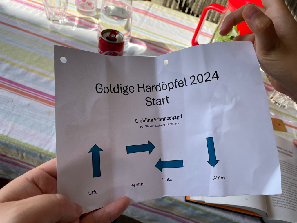 A close-up of the first instruction slip for the golden potato scavenger hunt. The text on the slip reads in Swiss-German: Goldige Härdöpfel 2024 (newline) Start. Then in a smaller font: E chline Schnitzeljagd (newline) PS. Alle Zettel wieder mitbringen. Translation: Golden Potato 2024 / Start / A small scavenger hunt. PS bring back all instructions slips. Below the text are four arrows pointing in the for cardinal directions with labels for each direction.