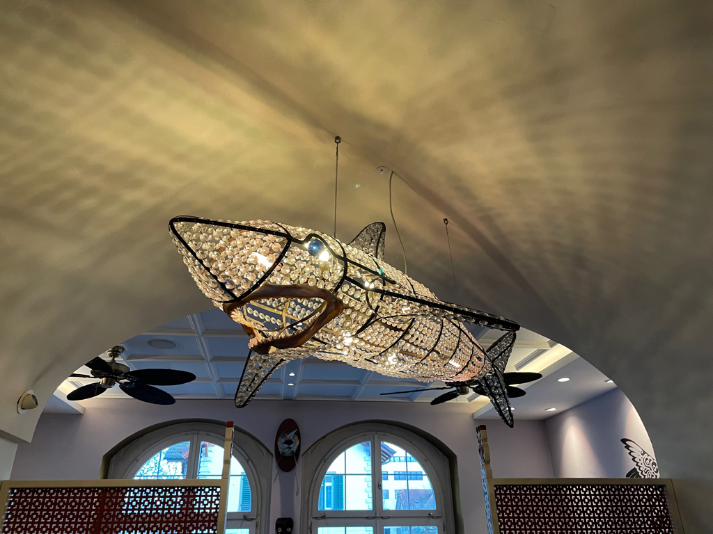 The image shows a unique chandelier in the shape of a large fish, possibly a marlin or shark. The fish's body is constructed from a framework of black metal, filled with numerous transparent crystal-like beads that give it a sparkling, luminous appearance. The chandelier emits light from within, enhancing the shimmer of the beads. The fish's eyes are made of blue, rounded accents, and its mouth is slightly open with a curved design.