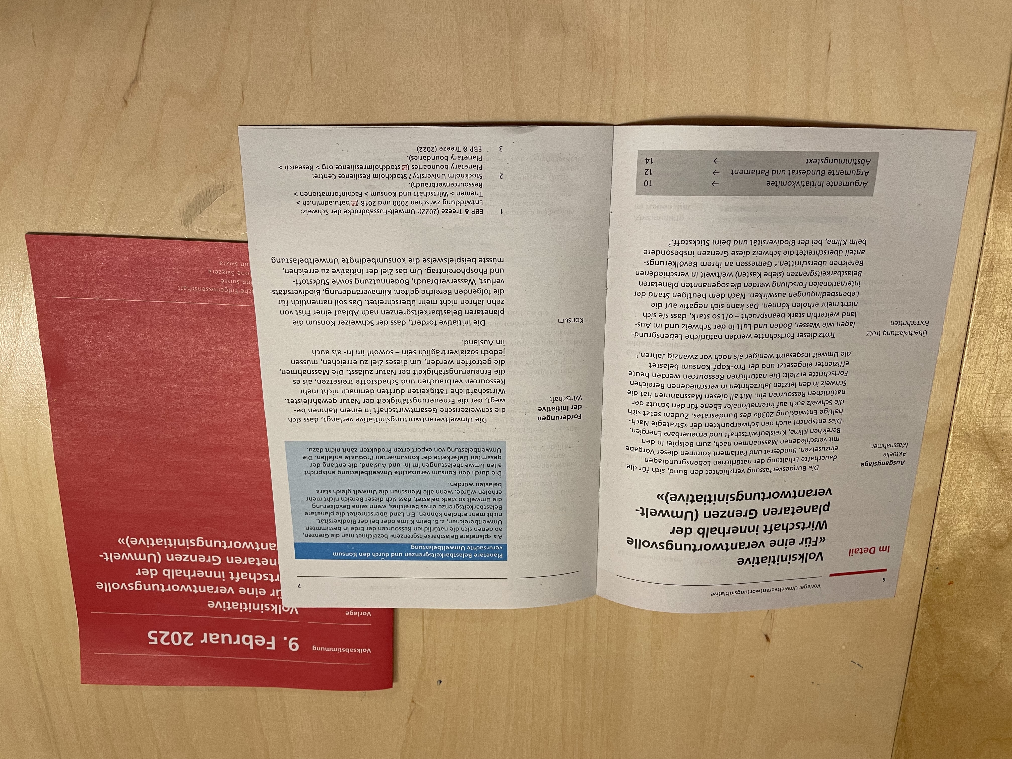 The image features the same booklet in two forms: one open and one closed. The booklet is printed in German and has a structured layout with headings, paragraphs, and highlighted sections. The left page has a bolded title and a red sidebar with labels, while the right page contains a blue-highlighted box. The red booklet on the right side, partially visible, has white text and a date—February 9, 2025—indicating it relates to an upcoming referendum. The overall design is clean and formal, with a focus on readability.