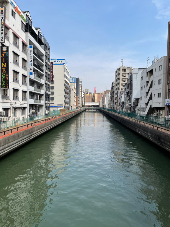 A canal flanked by buildings on both sides under a clear blue sky. The water is calm, reflecting the structures along its banks. Various signs in Japanese are visible on the buildings, which are mostly mid-rise and tightly packed. A bridge is seen in the distance spanning the canal.