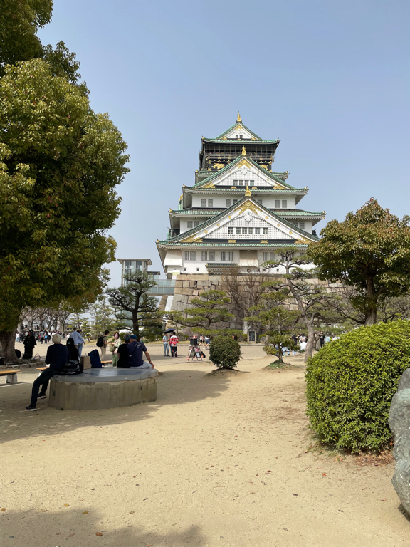A traditional Japanese castle with multiple tiers and green-tiled roofs stands prominently under a clear sky. The foreground features a sandy path lined with trees and shrubs. Several people are sitting on benches or walking around, enjoying the pleasant day.