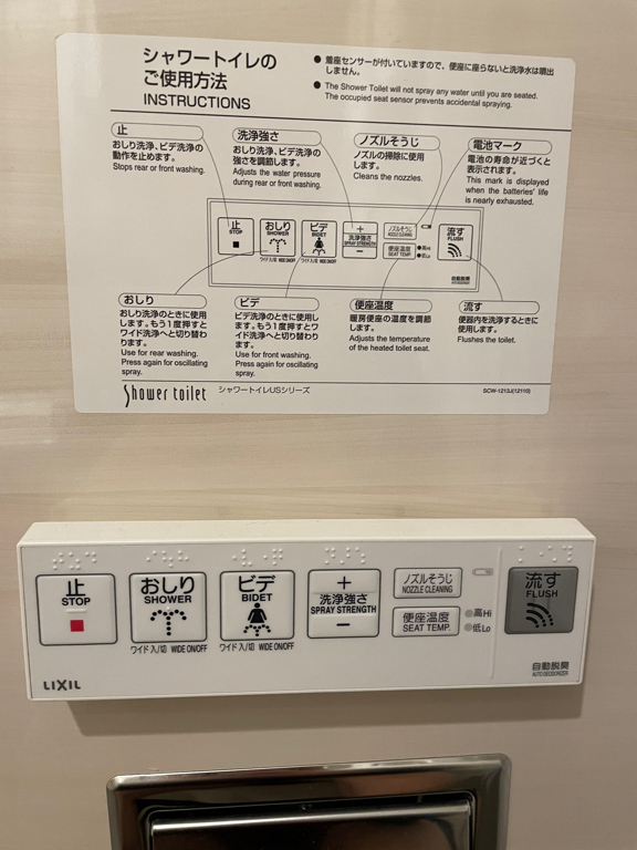 The image shows a control panel and instruction chart for a Japanese shower toilet. The control panel features buttons labeled with functions such as "Stop," "Shower," "Bidet," "Spray Strength," "Nozzle Cleaning," "Seat Temp," and "Flush." Each button has text in both Japanese and English, along with braille. Above the panel, an instruction chart provides detailed usage instructions for the toilet, including diagrams and descriptions in both Japanese and English. The brand name "LIXIL”